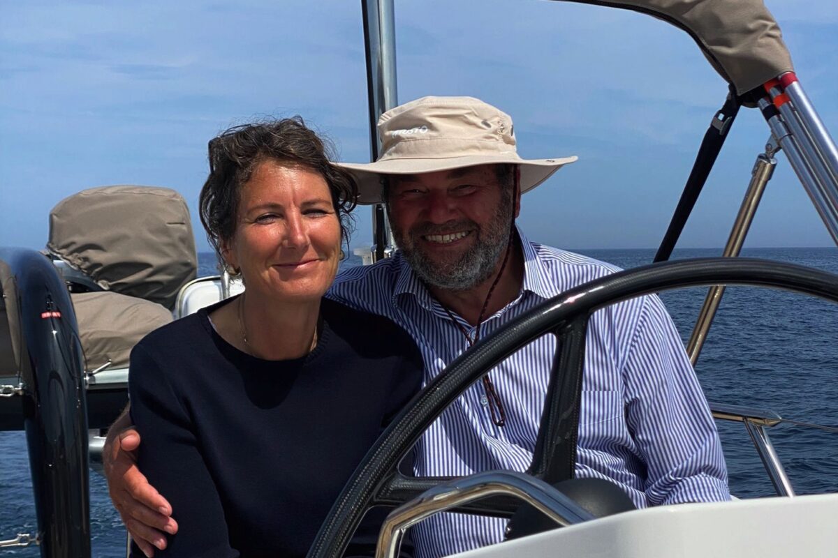 woman with dark hair and blue jumper and man with blue shirt and white hat on back of sailing yacht